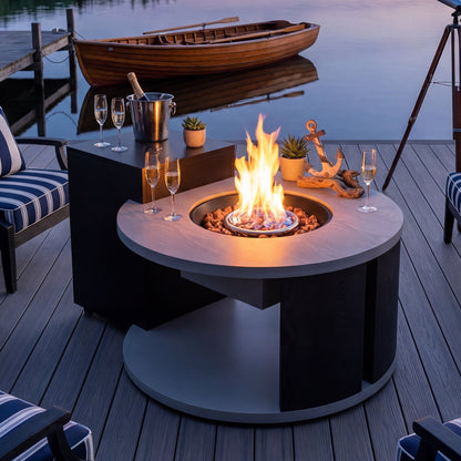 Fire pit table with flames on a wooden deck by a body of water, with chairs and decorative items around.